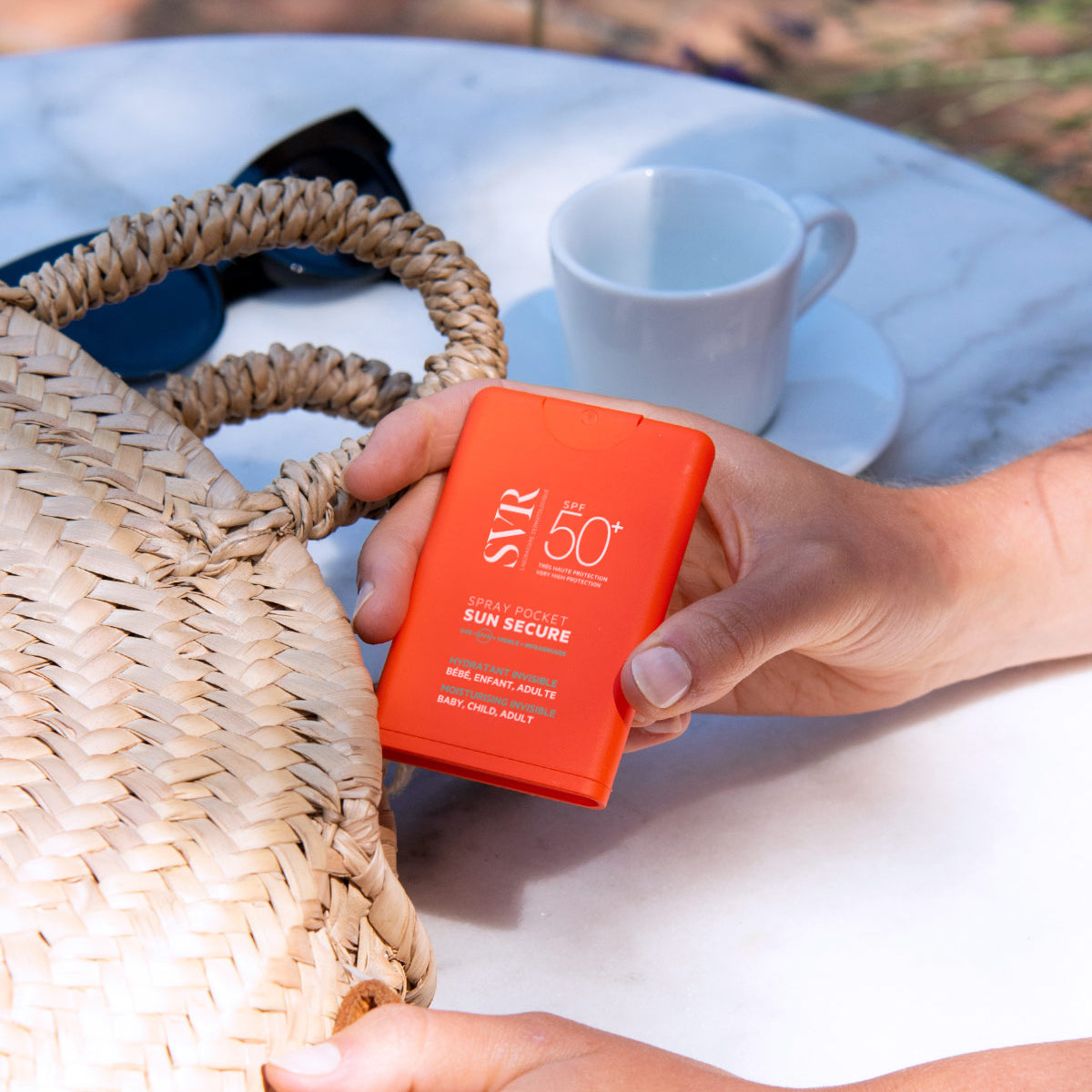 Spray solaire SVR Sun Secure SPF50+ format poche orange tenu en main près d'un sac en paille sur une table de terrasse.