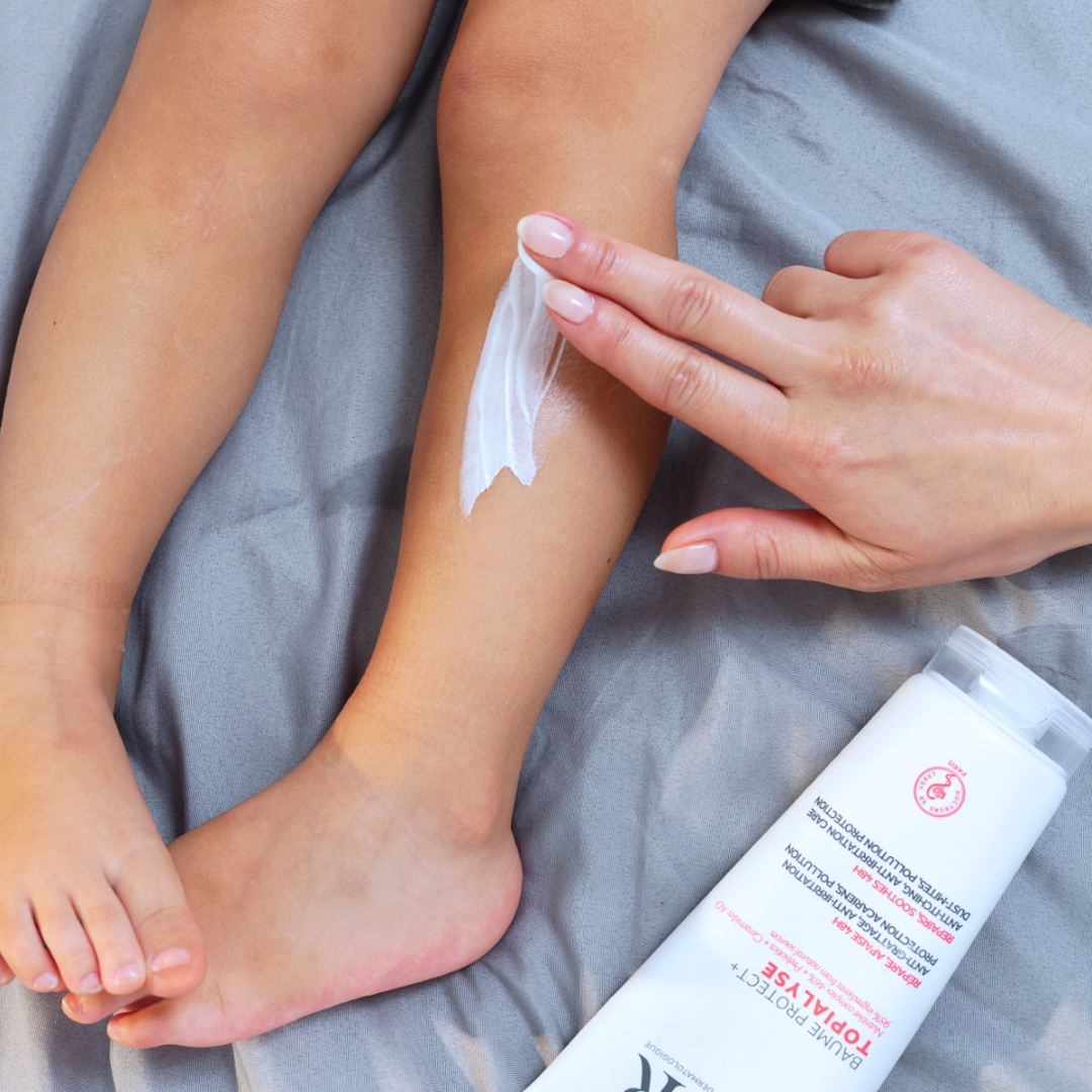 Main appliquant le baume réparateur TOPIALYSE Protect+ sur la jambe d'un enfant sur un drap gris.