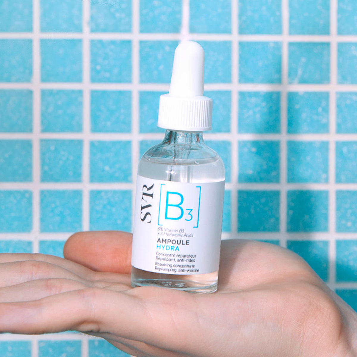 Flacon en verre transparent SVR Ampoule Hydra avec pipette blanche, tenu dans une main devant un carrelage bleu.
