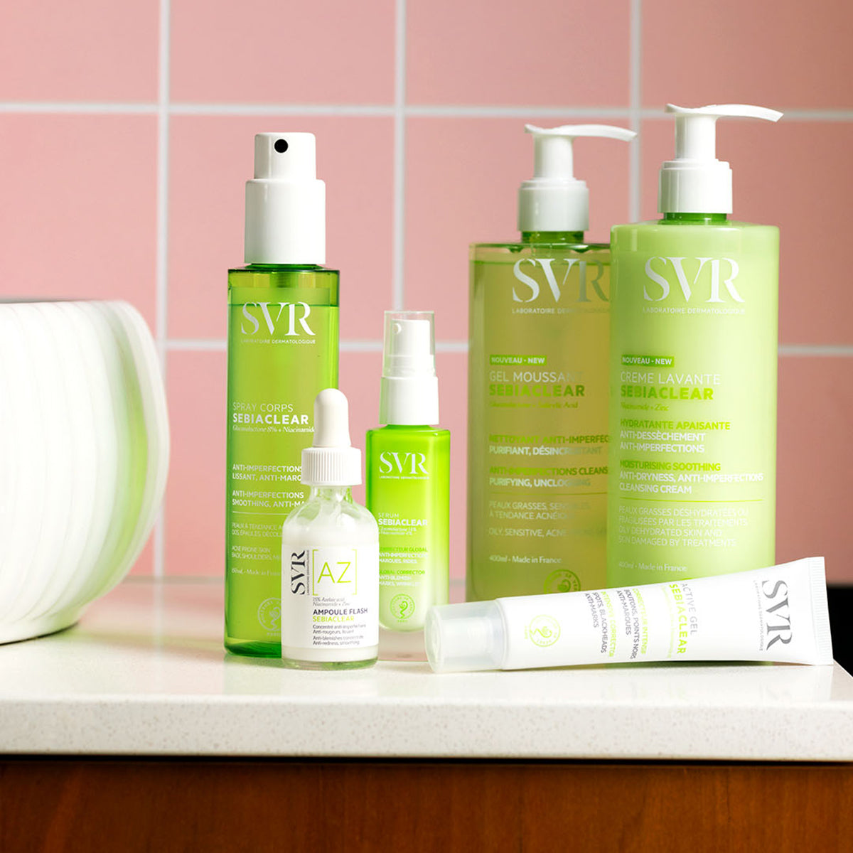 Gamme de soins SVR Sebiaclear : spray, sérum, gel moussant, crème lavante et tube de gel sur un comptoir devant un carrelage rose.