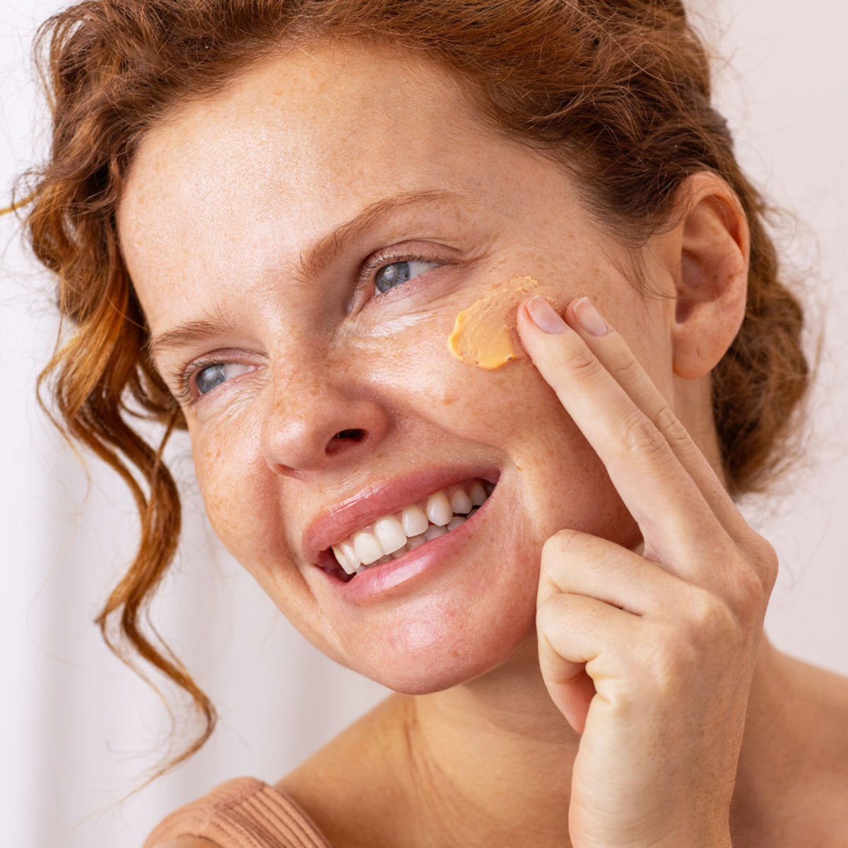Femme rousse souriante appliquant une crème solaire mousse abricotée SUN SECURE Blur SPF50+ sur sa joue.