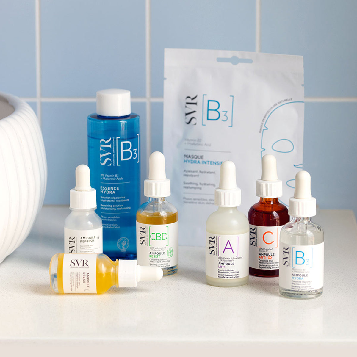 Masque Hydra Intensif SVR en biocellulose entouré de sérums Ampoule colorés sur un comptoir devant un carrelage bleu.