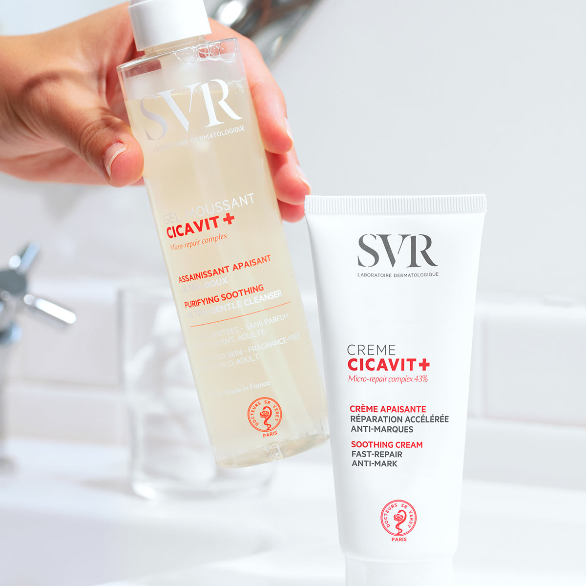 Tube de crème mains Cicavit+ SVR blanc et flacon de gel moussant transparent tenus en main devant un lavabo.