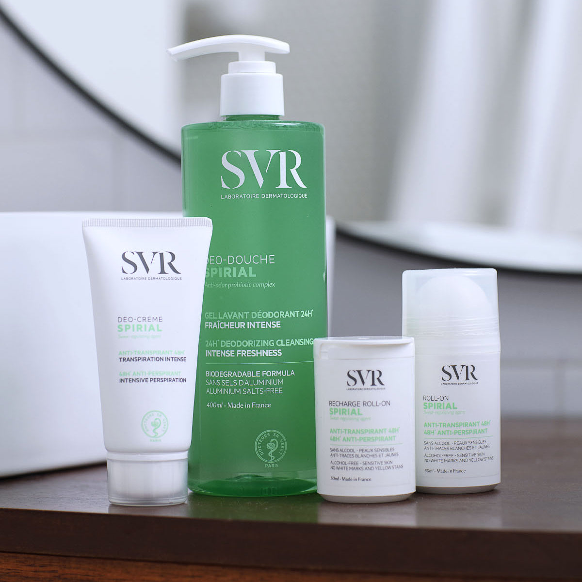 Gamme SVR Spirial : tube de crème, flacon pompe vert de gel douche et deux déodorants roll-on sur un support en bois.