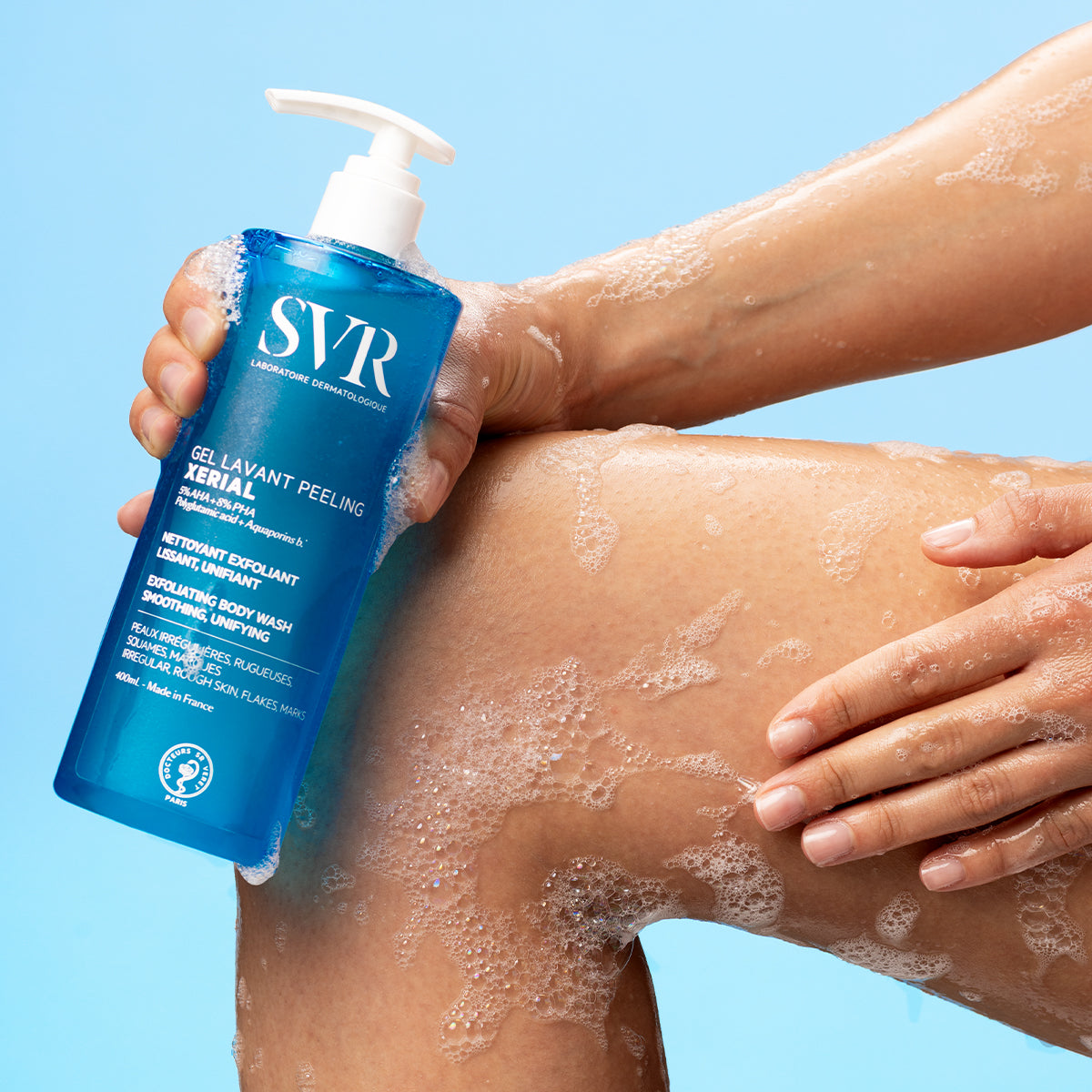 SVR XERIAL Gel Lavant Peeling bottle held against a soapy leg, featuring a blue pump bottle with white text on a blue background.