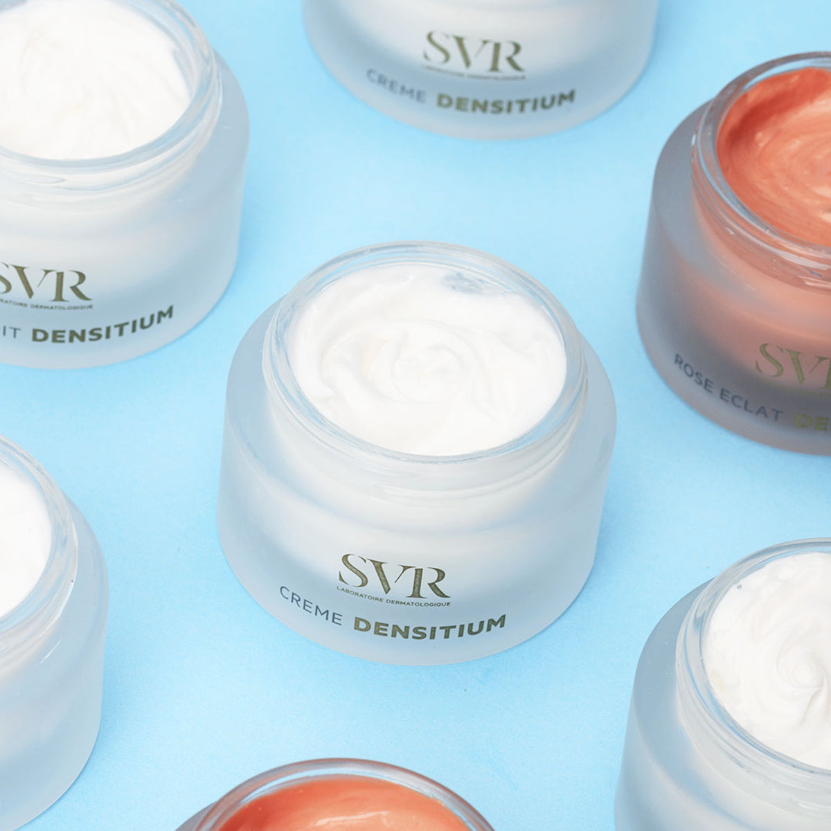 Open jars of SVR Densitium creams with white and rose-tinted formulas in frosted glass containers on a light blue background.