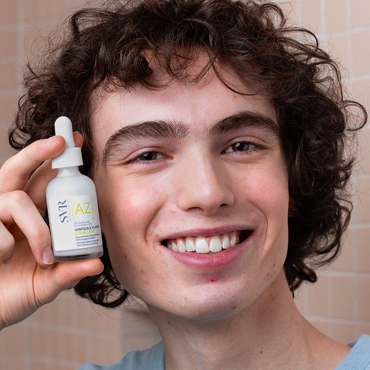 Jeune homme souriant tenant un flacon compte-gouttes blanc SVR Sebiaclear Ampoule Flash contre un mur carrelé.