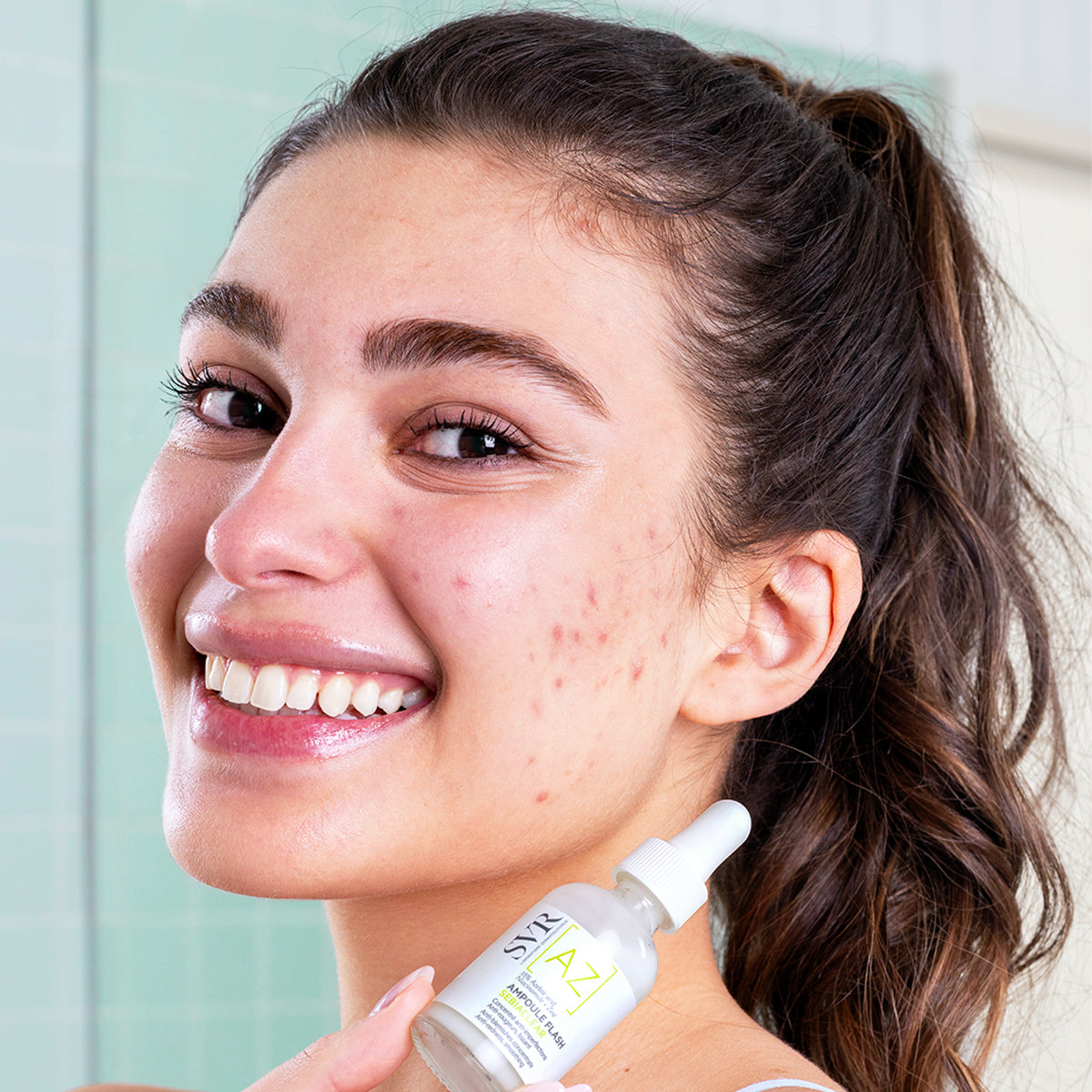 Smiling woman holding a white SVR Sebiaclear Ampoule Flash bottle with a dropper cap in a bright bathroom setting.
