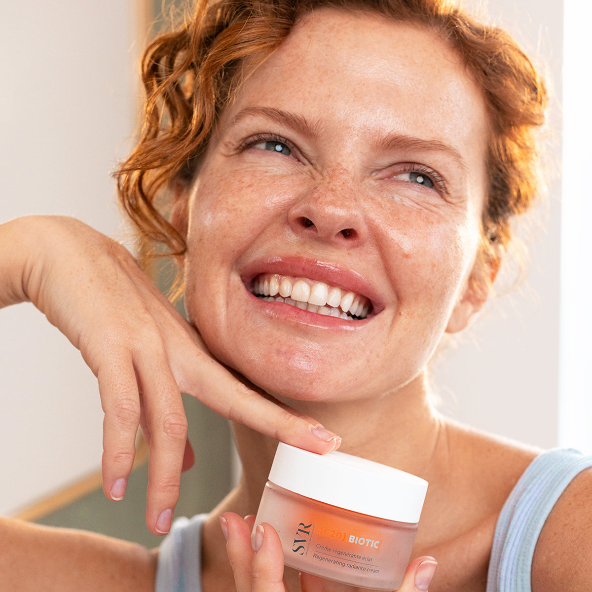 Smiling woman with freckles holding a jar of SVR C20 Biotic regenerating radiance cream with a white lid.