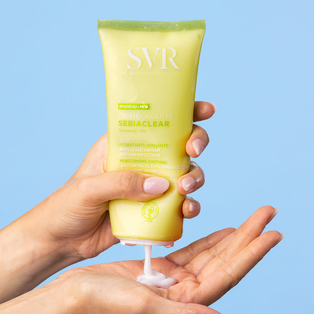 Tube de crème lavante SVR Sebiaclear jaune pâle pressé dans une main, délivrant une texture onctueuse sur fond bleu.