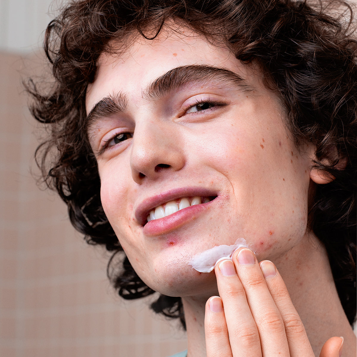 Young man with curly hair applying white CICAVIT+ Crème HPPI to a blemish on his chin while smiling at the camera.