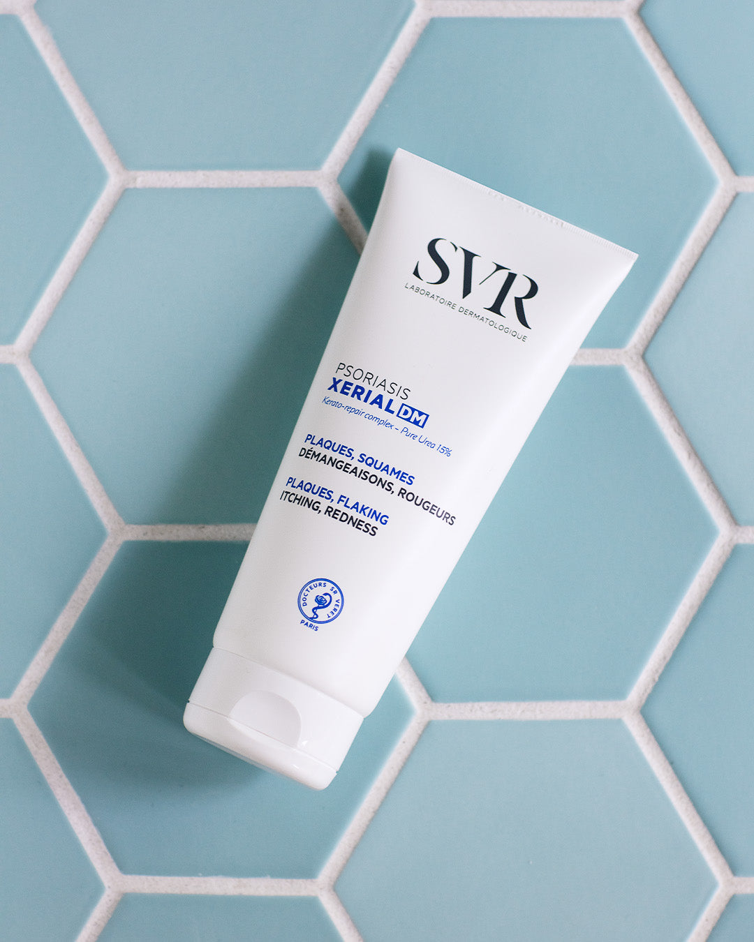 White SVR XERIAL DM Psoriasis cream tube with blue text on a light blue hexagonal tiled background.