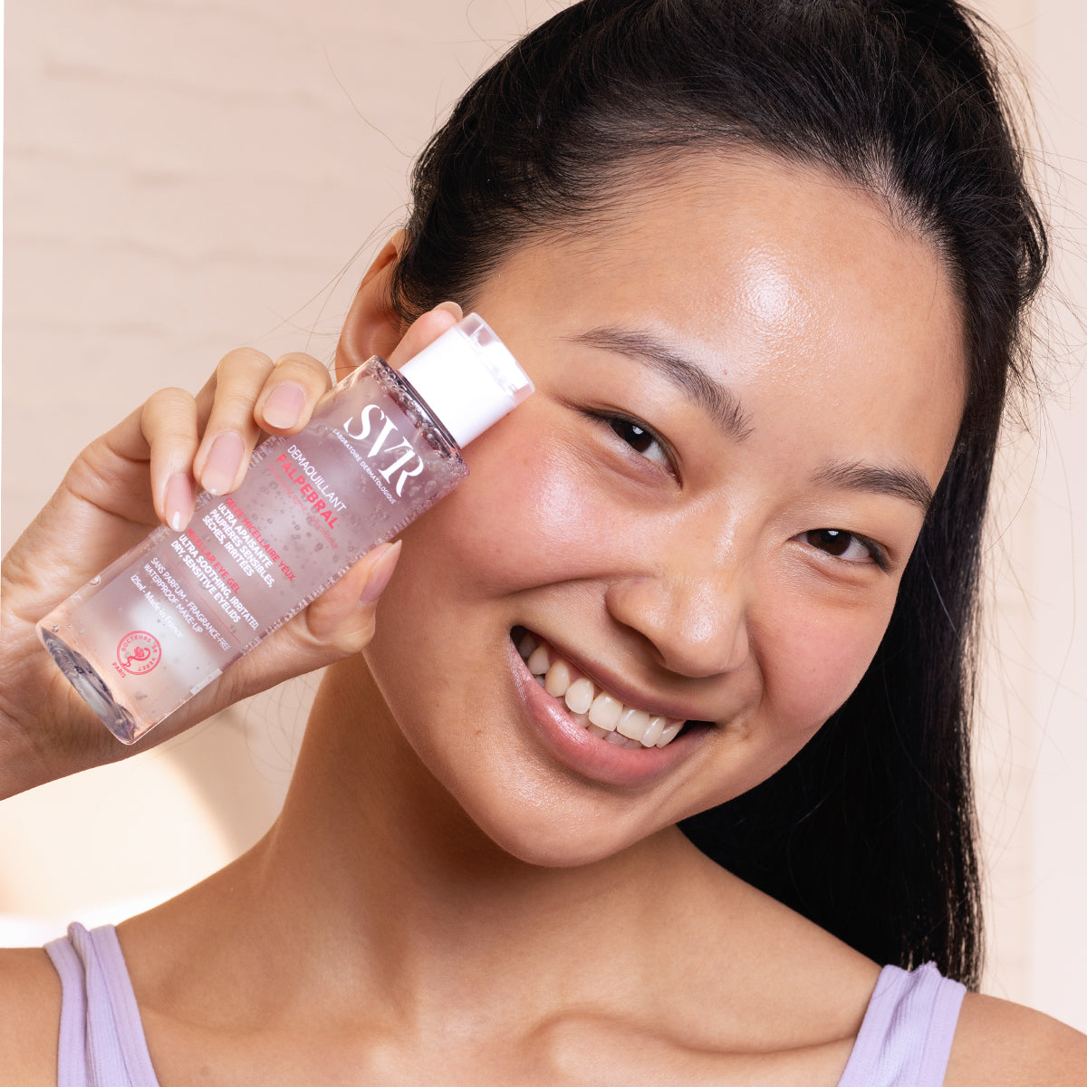 Femme souriante tenant un flacon transparent de démaquillant SVR Palpebral à la texture gel pour yeux sensibles.