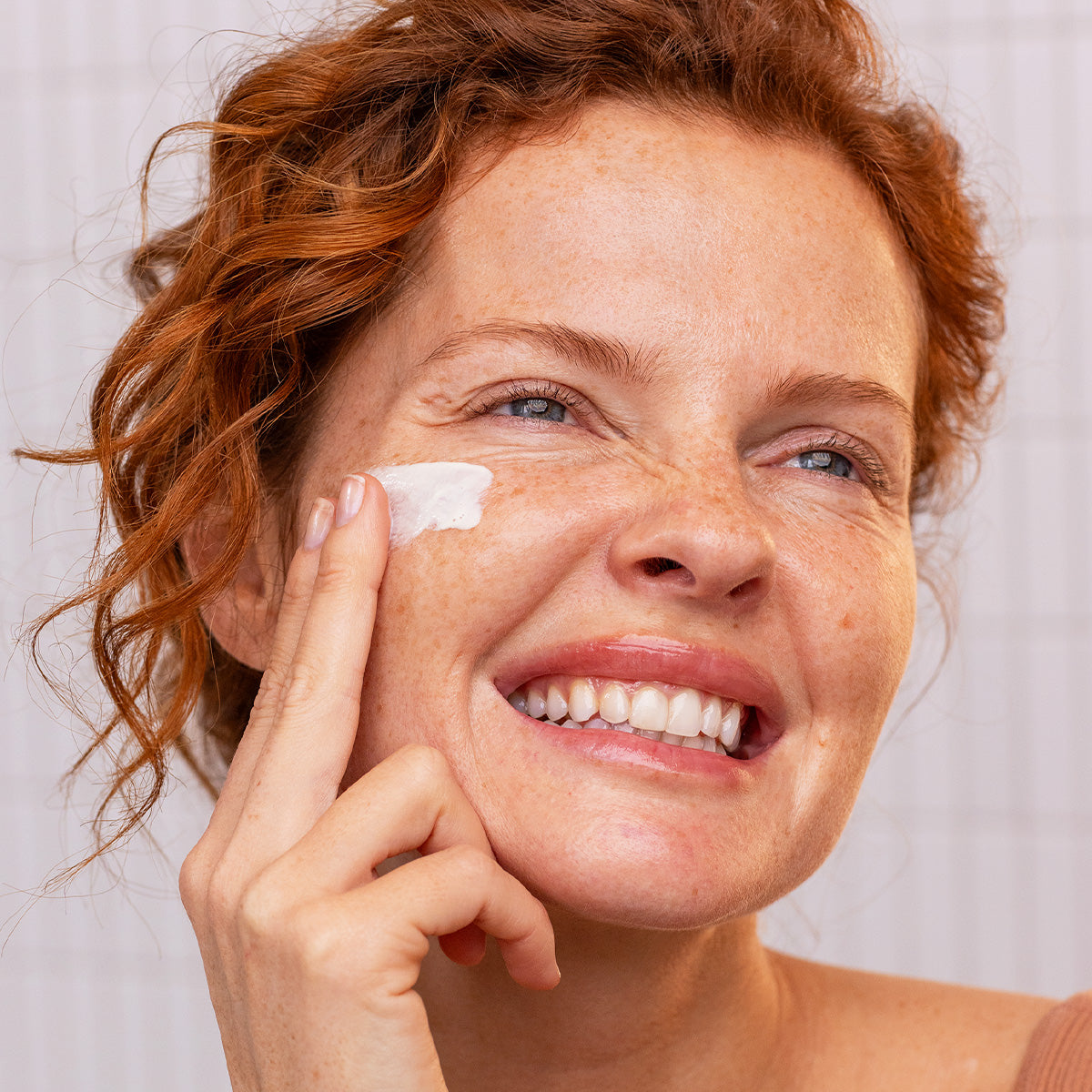 Smiling woman with red hair applying white SUN SECURE SPF50+ fluid sunscreen to her cheek.