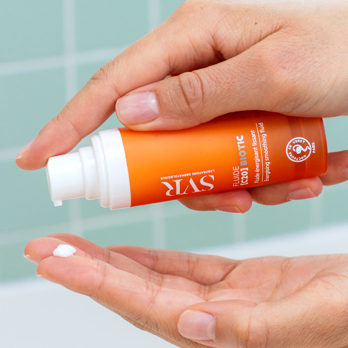 Hand holding an orange SVR Fluide [C20] BIOTIC pump bottle, dispensing a small drop of white smoothing cream into the palm.
