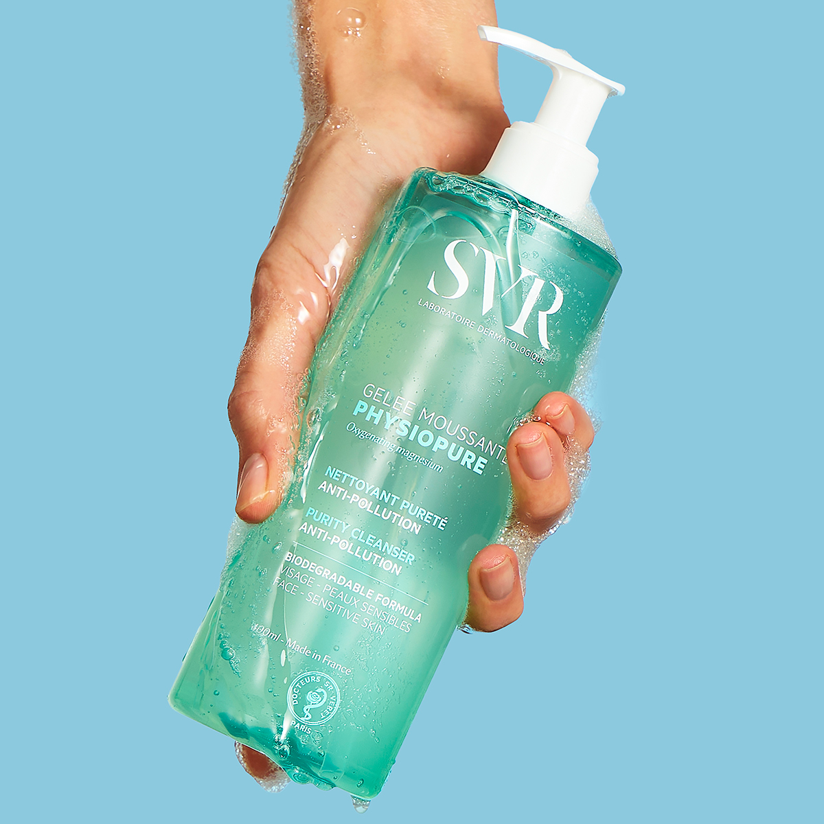 Main tenue en main d'un flacon pompe SVR Physiopure Gelée Moussante bleu translucide avec une mousse nettoyante légère.