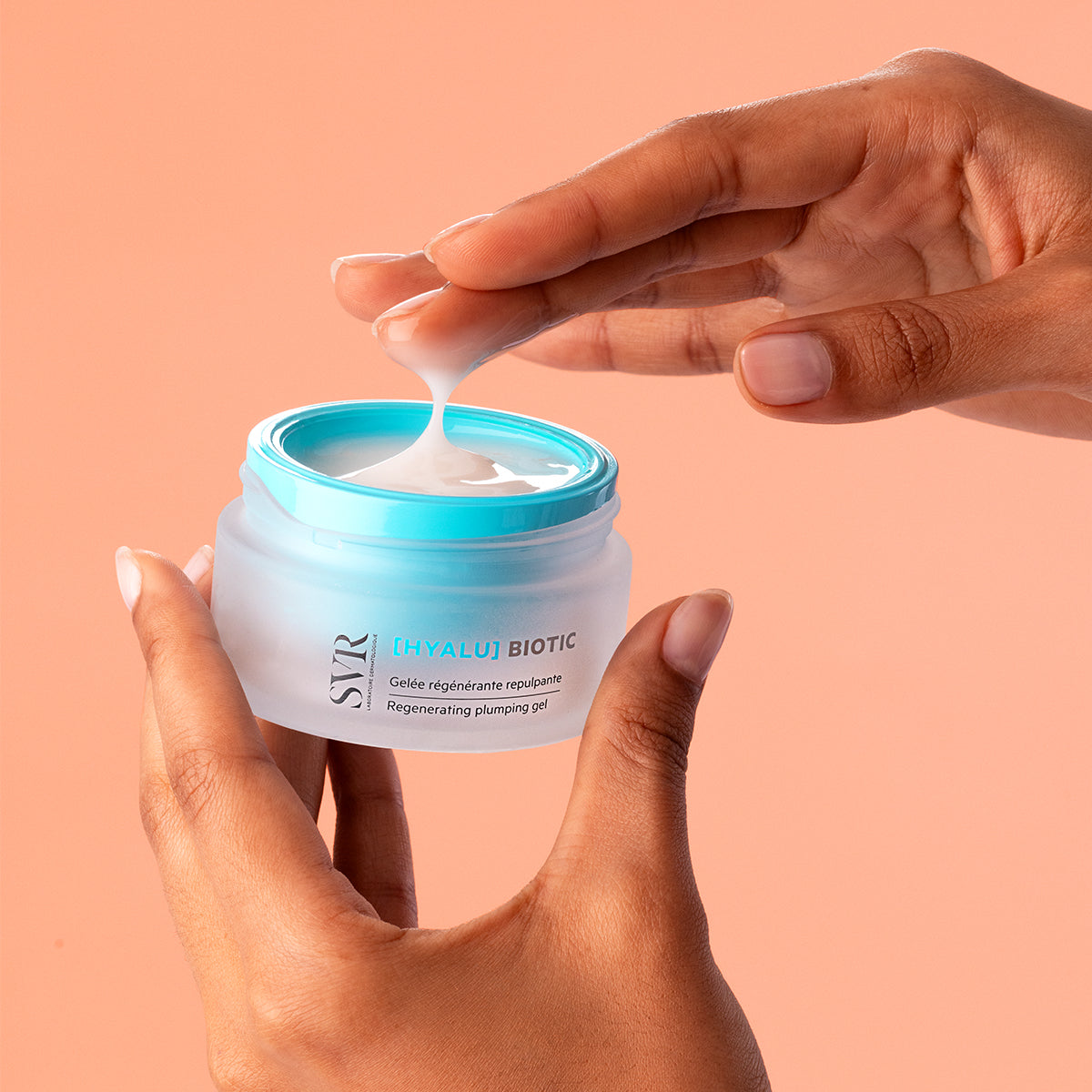 Hands holding a frosted glass jar of SVR [HYALU]Biotic regenerating plumping gel with a bright blue lid.