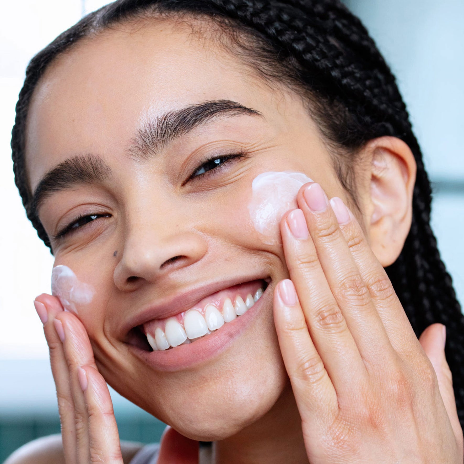 Femme souriante appliquant une crème hydratante blanche sur ses joues pour sa routine solaire quotidienne.