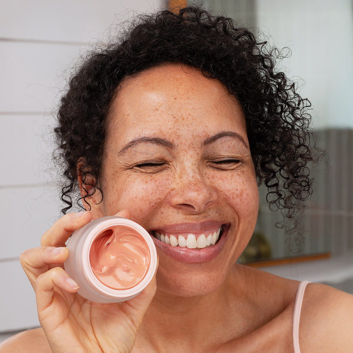 Smiling woman holding an open jar of Densitium Rose Eclat cream with a soft pink, creamy texture.