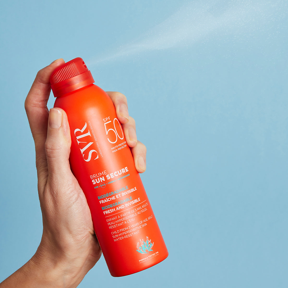 Main tenue à la main vaporisant la brume solaire SVR Sun Secure SPF50 dans un flacon orange vif sur fond bleu.