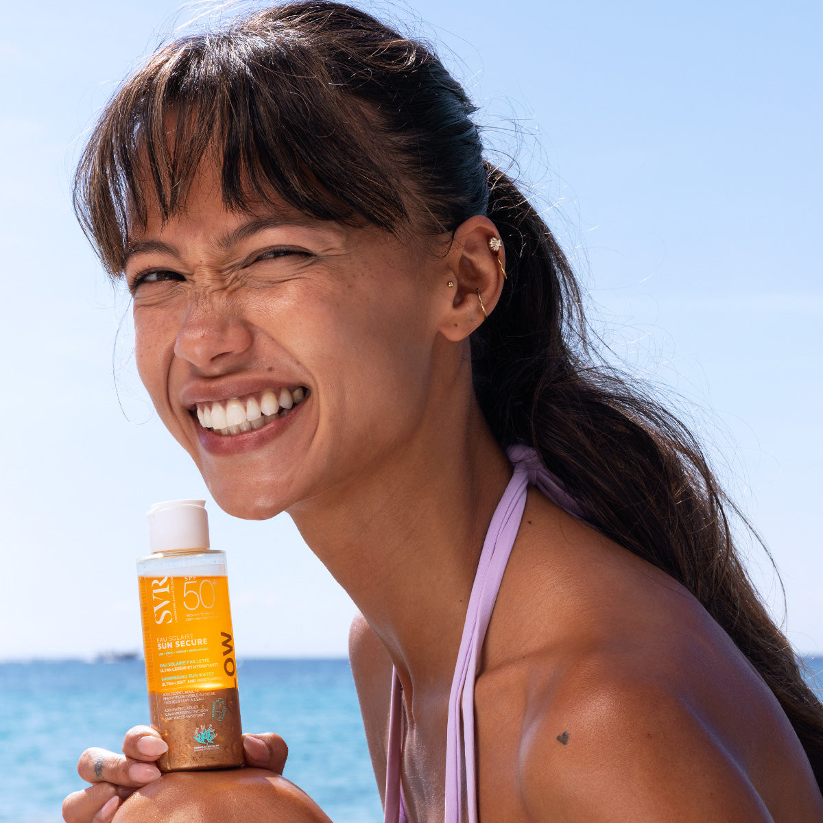 Smiling woman holding a bottle of SVR SUN SECURE Eau Solaire Glow SPF50+ against a sunny beach background.