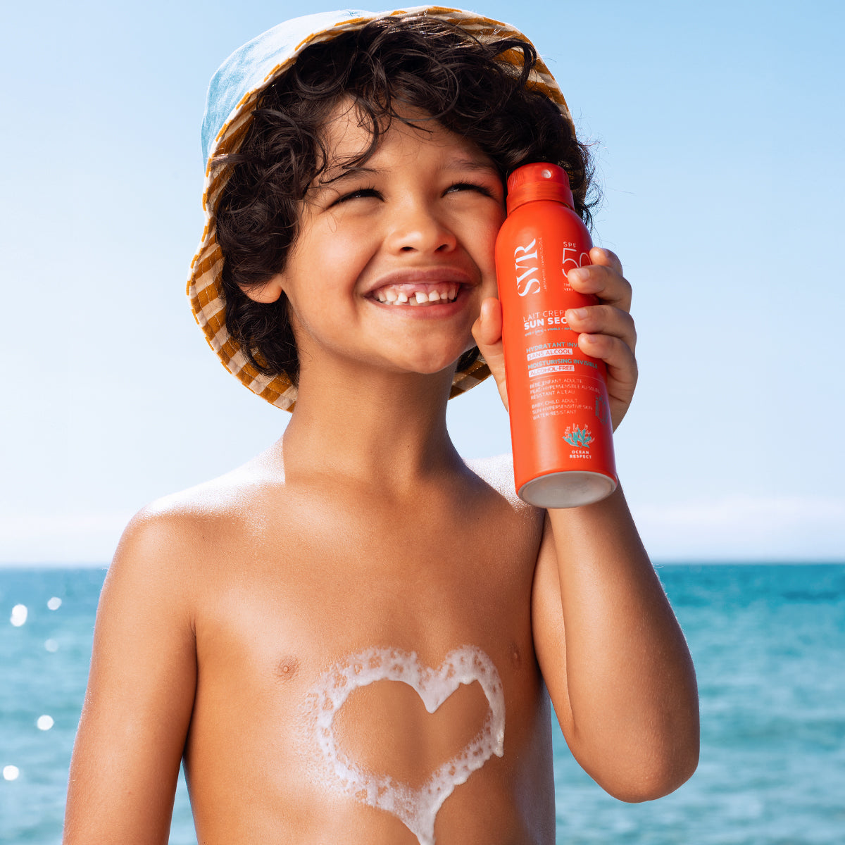 Enfant souriant avec un chapeau tenant un spray solaire SVR Sun Secure SPF50+ et un cœur en mousse sur la poitrine à la plage.