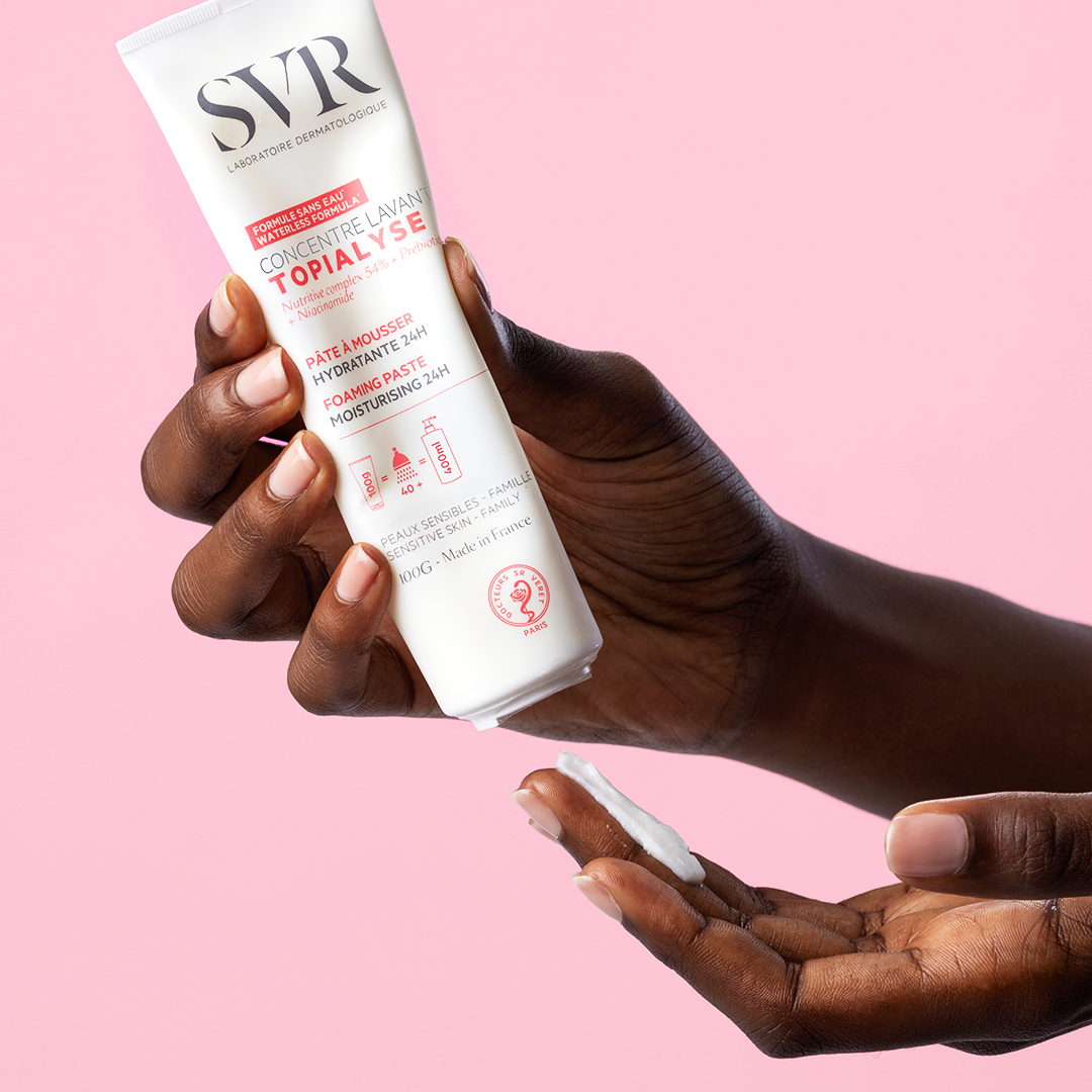 Hand holding a 100g white tube of SVR Topialyse Concentré Lavant, dispensing a small amount of white foaming paste.