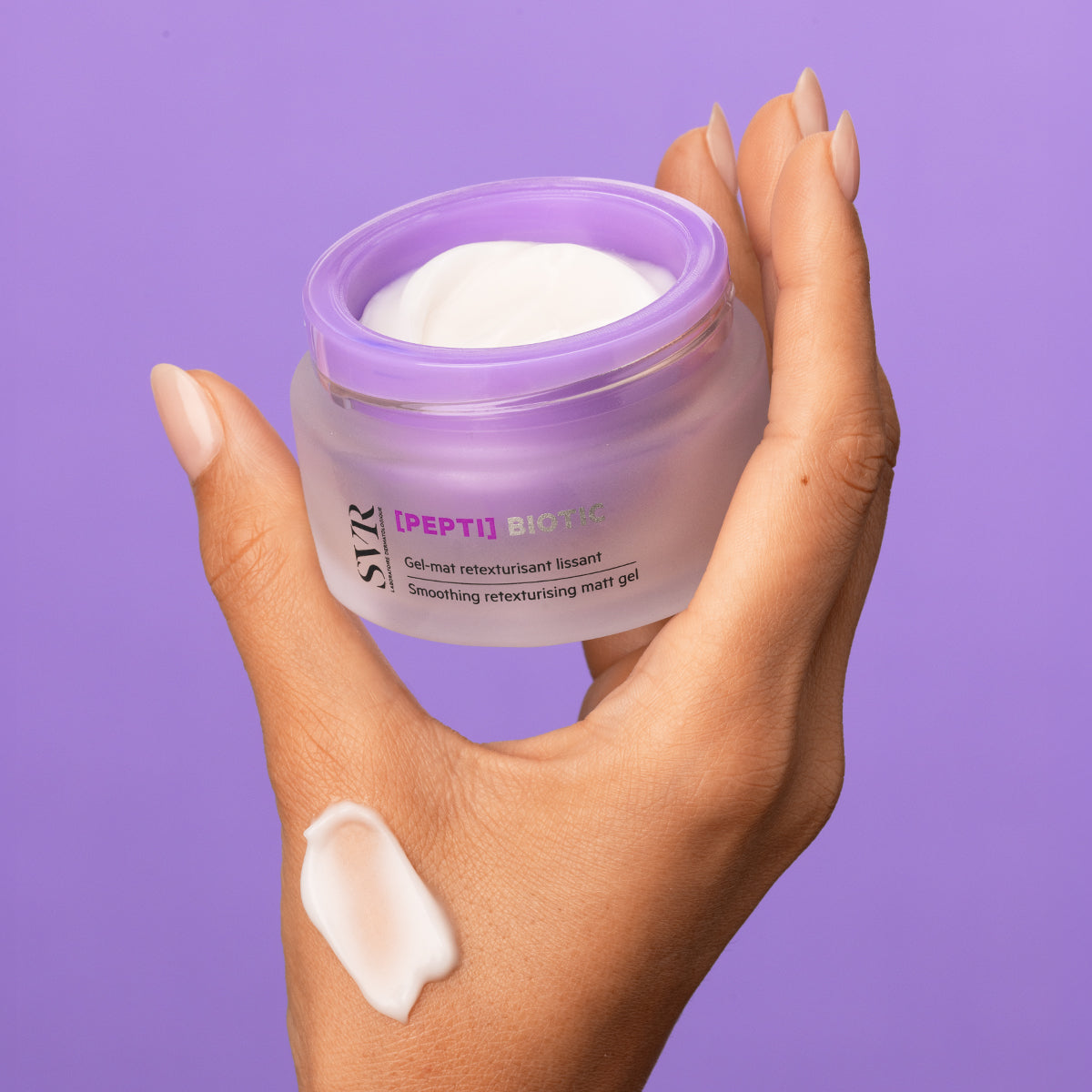 Hand holding a frosted glass jar of SVR [PEPTI]Biotic smoothing matt gel with a white cream swatch on the skin.