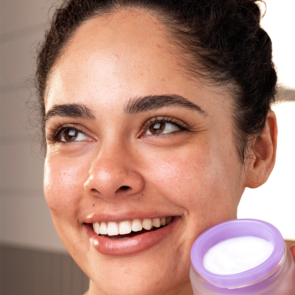 Smiling woman with glowing skin holding an open jar of white PEPTI Biotic cream with a lavender lid.