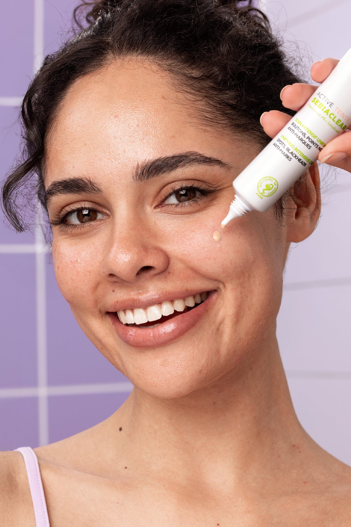 Smiling woman applying SVR Sebiaclear Active Teinte tinted cream to her cheek against a purple tiled background.