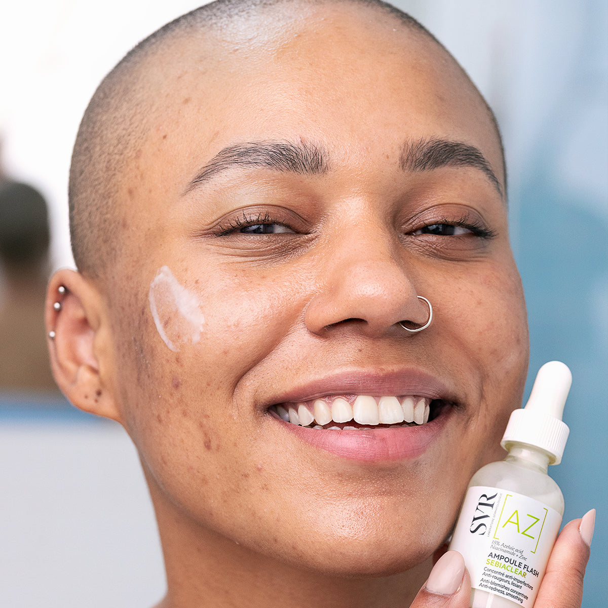 Personne souriante tenant un flacon compte-gouttes SVR Sebiaclear Ampoule Flash avec une texture lactée appliquée sur la joue.