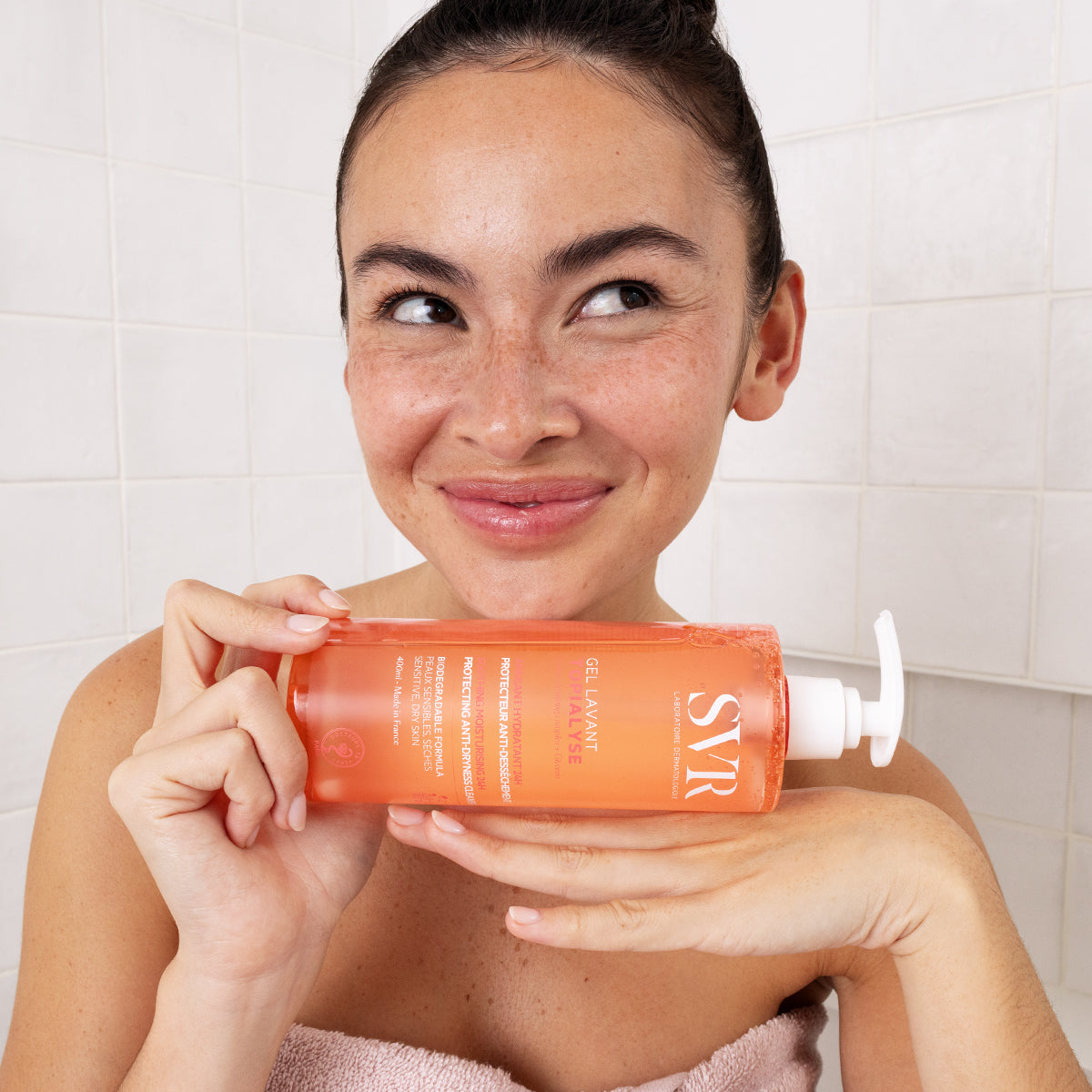 Femme souriante tenant un flacon pompe orange de gel lavant SVR Topialyse devant un carrelage blanc.