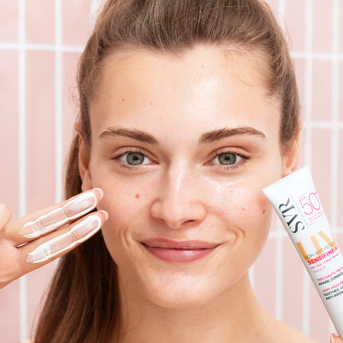 Femme souriante appliquant la crème SVR Sensifine AR SPF50+ sur son visage devant un carrelage rose.